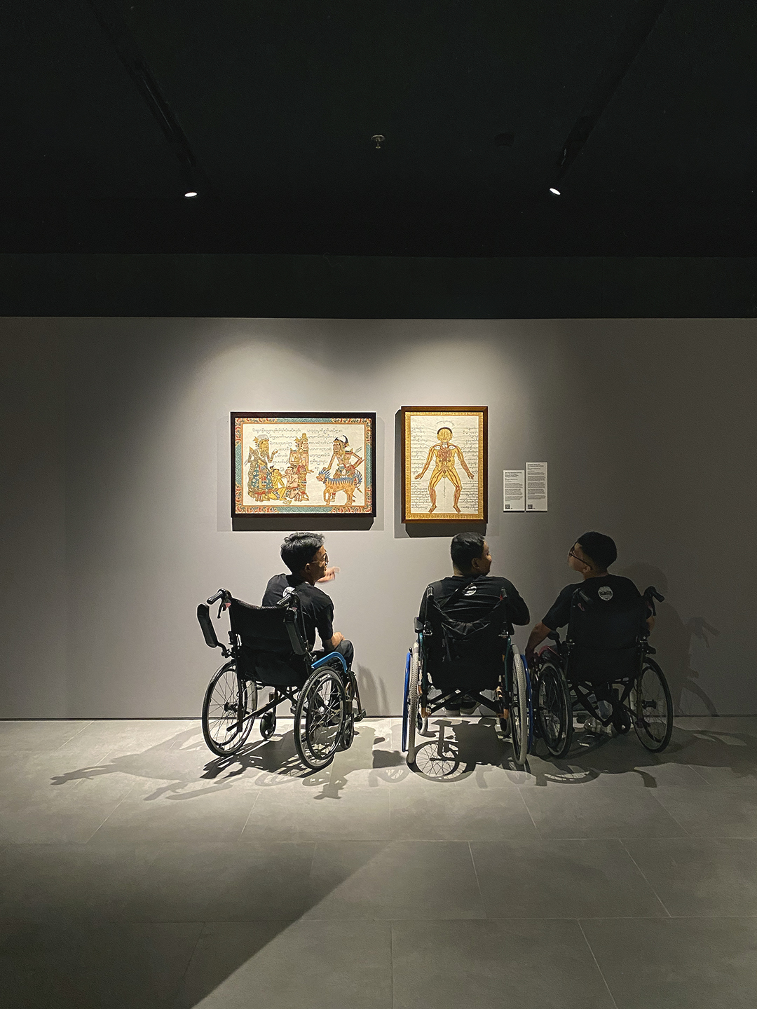 Three people in wheelchairs viewing framed artwork in a dimly lit gallery.